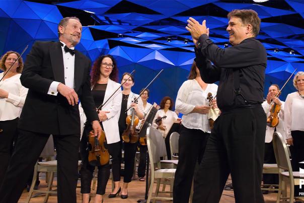 The camera has an on-stage view of an orchestra. All the musicians are dressed in white tops and black pants, holding their instruments. In front of them are two male conductors (?) approaching each other while clapping for their performers.