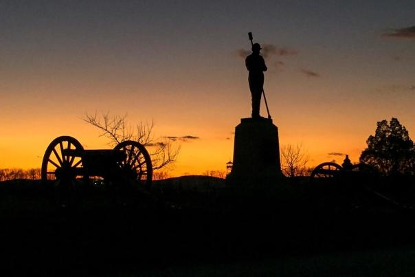 Silhouettes of a statue and a cannon on a field with a sunset in the background