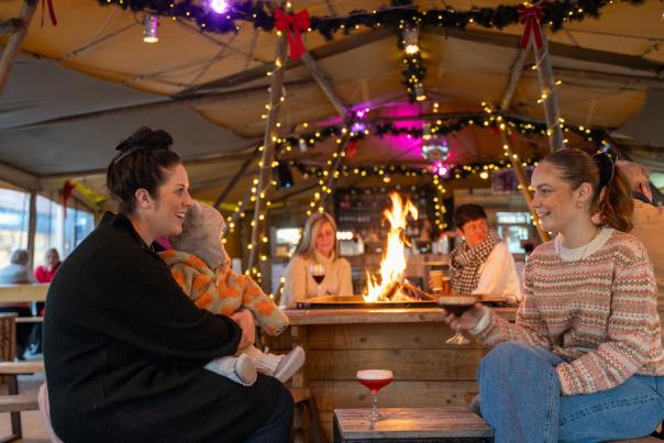 two people and a baby sitting in a bell tent with fairy lights and an open fire enjoying a cocktail