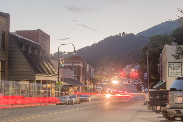 Red lights from cars stream through the photo on a road, the road is lined with old buildings. There are some cars parked on either side of the street and a large mountain is seen in the background.