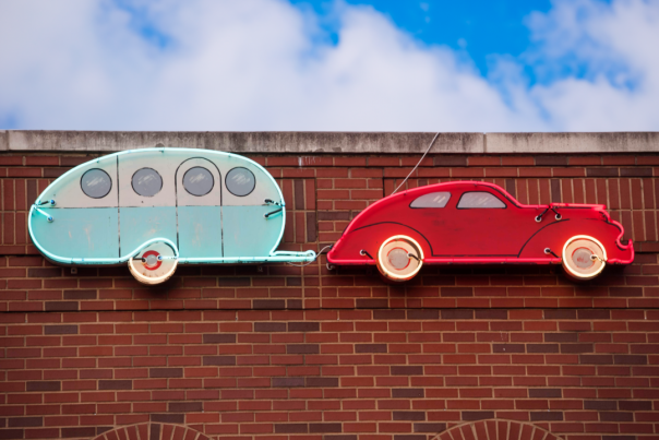 Neon sign of a red vintage car towing a light blue camper, mounted on a brick wall against a blue sky.