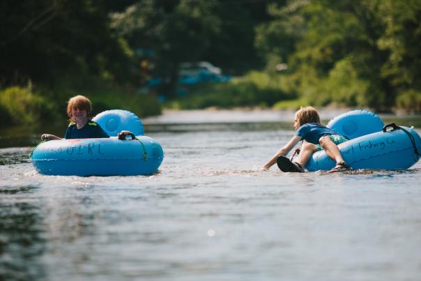 Two children are seen in blue river tubes floating on the river. One smiles big for the camera while the other leans over their tube to splash the other.