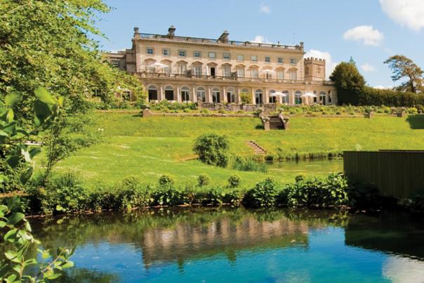 A lake in the foreground with reflections of the sky, with a green hill in the background leading up to Cowley Manor, a large country house.