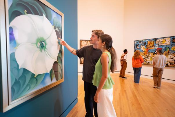 A couple admires a large floral painting in an art gallery, surrounded by other visitors exploring various artworks. Bright, spacious interior.