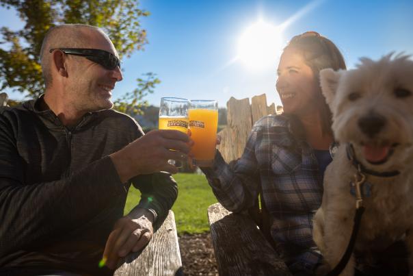 A white man and white woman cheers two beer glasses from Wisconsin Brewing Company outside while holding a small white dog