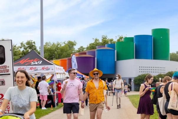 A vibrant outdoor scene with colorful buildings in the background. People stroll along a path, enjoying a lively event with tents and activities.
