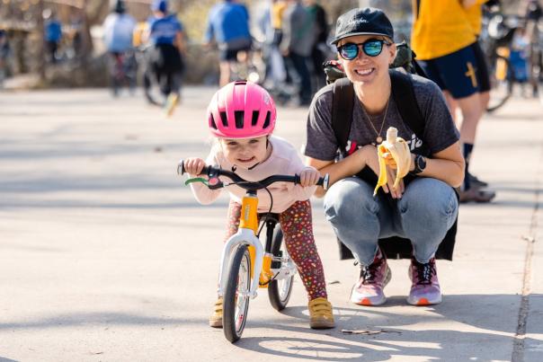 A child wearing a pink helmet learns to ride a balance bike with encouragement from an adult during the Collegiate MTB Championships, surrounded by cyclists.