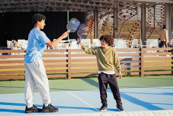 Two children play with paddles on a blue court, surrounded by wooden fencing and outdoor seating. Sunlight illuminates the scene.