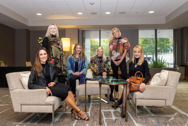 Six well-dressed women, 40s-60s, smile at the camera. Four of them are sitting on fancy white armchairs angled toward each other. The remaining two women stand between them.