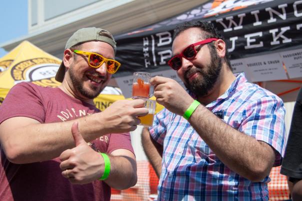 Two men wearing colorful sunglasses and green wristbands smile and clink their beer tasting glasses at an outdoor festival. One gives a thumbs up while the other raises his glass, with brewery tents and signage visible in the background.