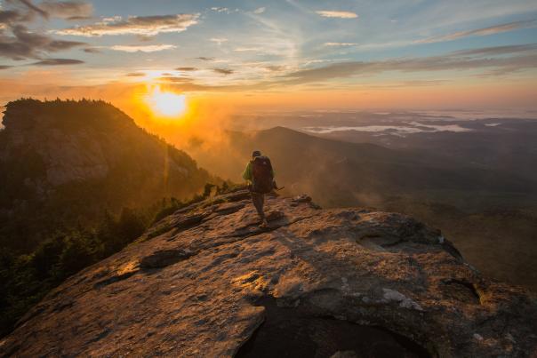 A hiker with a large backpack is walking on a large boulder into the sunrise with expansive vista views.
