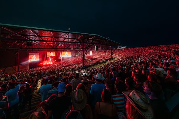An open-air concert stage with thousands of people watching a show illuminated in a red glow from the stage lights against the pitch black of the night sky.