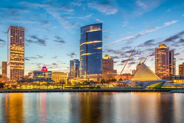 A sunset view of downtown Milwaukee reflecting on Lake Michigan, featuring the illuminated Milwaukee Art Museum, modern high-rise buildings, and a colorful sky with scattered clouds.
