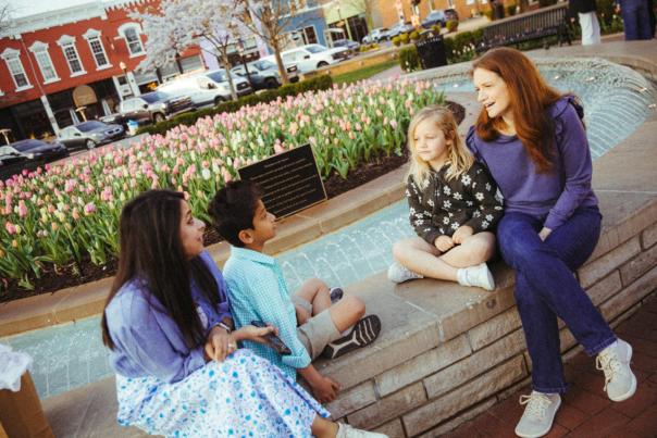 A woman with long auburn hair sits on a brick ledge next to a young girl with blonde hair in a patterned jacket. Across from them, a woman with long dark hair in a blue jacket and patterned skirt sits next to a young boy in a blue checkered shirt and khaki shorts. They appear to be talking near a fountain and a bed of pink tulips.