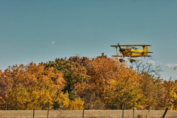 Yellow airplane flying above a treeline in the fall. Trees are yellow, orange, and green.