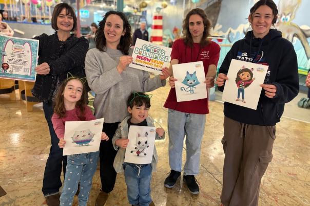 Six people in comfortable clothes (four adults in back, a young girl and boy in front) smile at the camera, each showing off a flier for the MEOWseum exhibit or a character portrait from the Gabby's Dollhouse animated TV show.