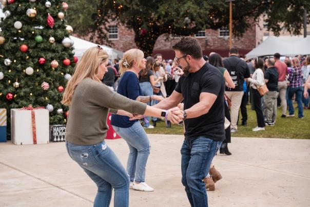 woman in green sweater and jeans dances with man in black shirt and jeans in front of a christmas tree downtown at the annual smoke and barrel festival