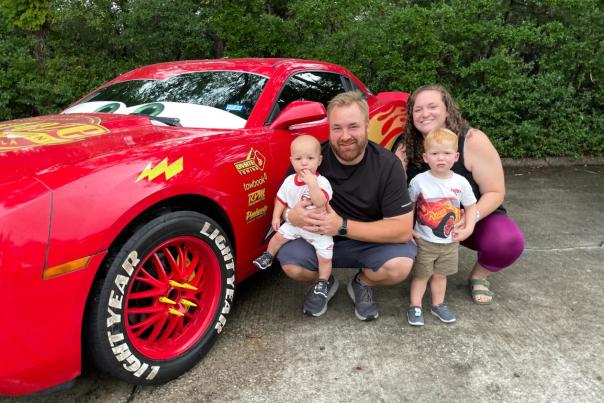 A young family of four squats beside a life-sized model of Lightning McQueen from Disney Pixar's "Cars." The sons (maybe 1 and 3 years old) are wearing Lightning McQueen shirts and khaki shorts.