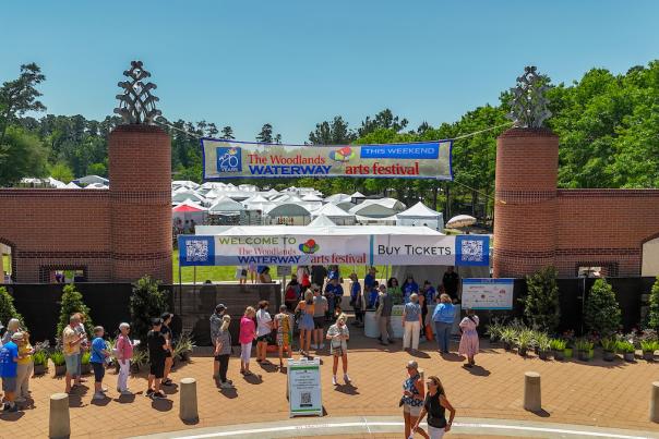 The entrance of Town Green Park and The Woodlands Waterway Arts Festival 2025 features two red-brick pillars topped with large metal pinecones. On the brick sidewalk in front of the gates, dozens of festival-goers mill about. Behind the gates are dozens of white tents featuring various kinds of fine art.