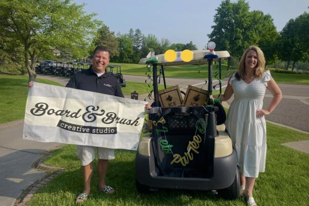 Board & Brush Woodbury Studio staff getting ready for Chamber golf tournament, holding a promotional sign next to a golf cart.