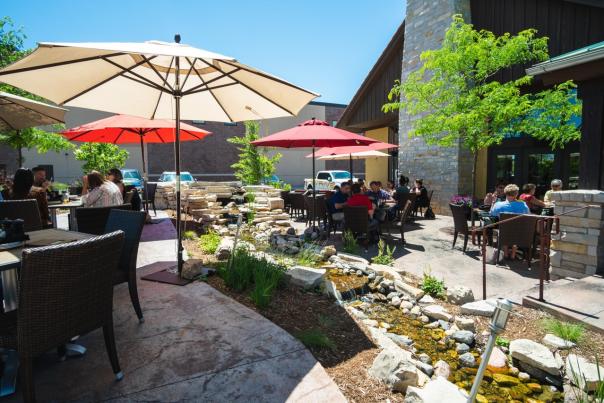 People dining at outdoor patio tables under umbrellas near a small garden and water feature on a sunny day.