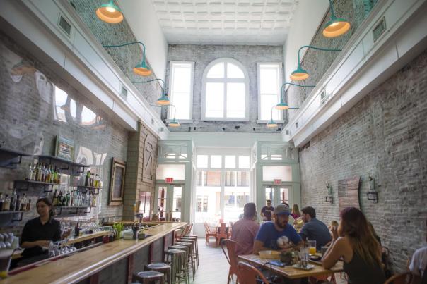 A restaurant with exposed brick walls and large windows with sunlight shining on people dining