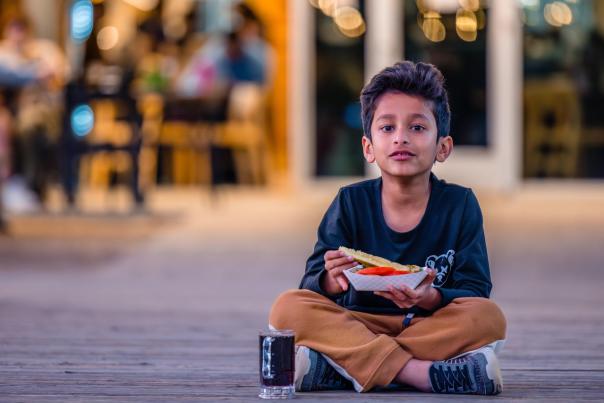 A young boy sits cross-legged, holding a plate with a hot dog, with a glass of soda beside him. A lively outdoor dining scene is blurred in the background.