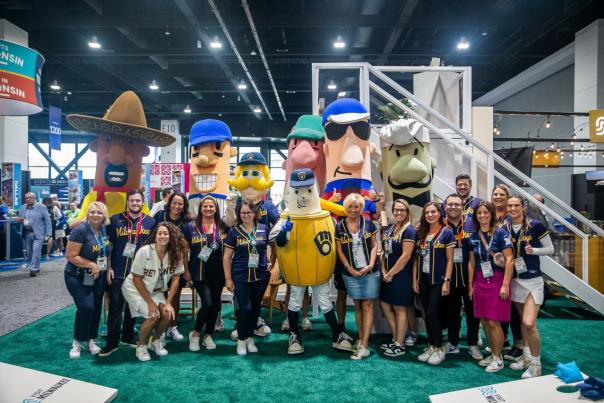 Group of people posing with the Milwaukee Brewers racing sausages and mascot at an indoor convention or expo booth, surrounded by signage and displays.