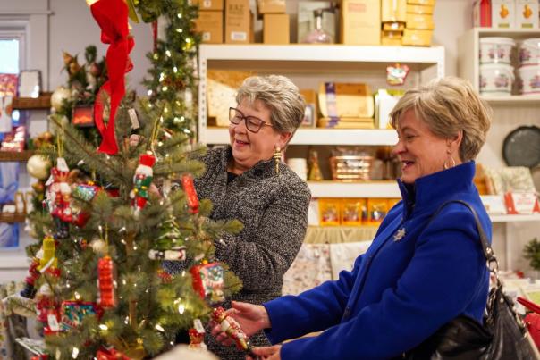 Two women decorate a Christmas tree with colorful ornaments in a cozy shop filled with holiday items. Shelves in the background display festive goods.