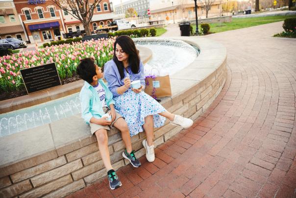 A woman and boy sit beside a fountain, near blooming tulips in a vibrant park setting, with shops in the background.