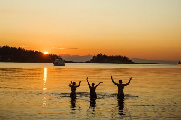 Three people standing in calm ocean water at sunset with arms raised.