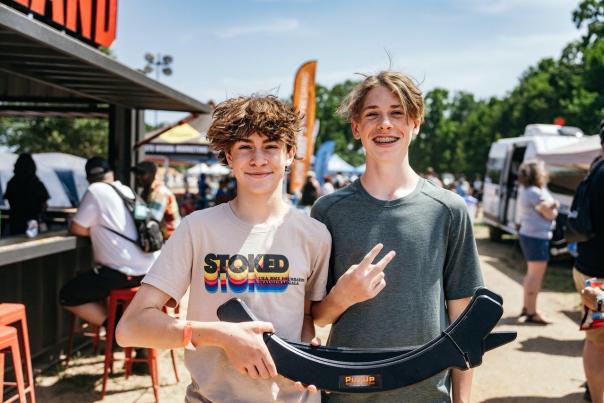 Two young men stand outdoors at a lively event, smiling and holding a curved object. Colorful banners and a crowd are visible in the background.