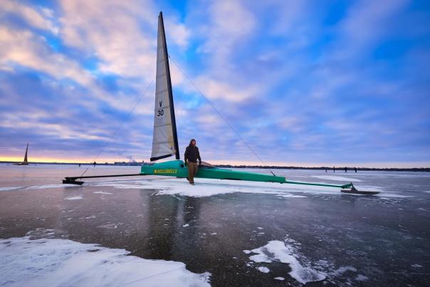 A sailboat sitting on top of the frozen Lake Monona. A man is standing next to the boat and the sun is setting