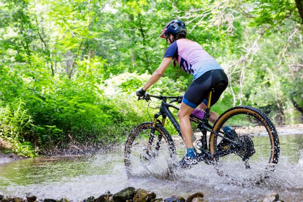 A person is riding a mountain bike through a shallow stream in a Coler Mountain Bike Preserve. The rider is wearing a helmet, a short-sleeved jersey with a gradient design, and cycling shorts. The bike is black with brown tires, and water is splashing as the bike moves through the stream.