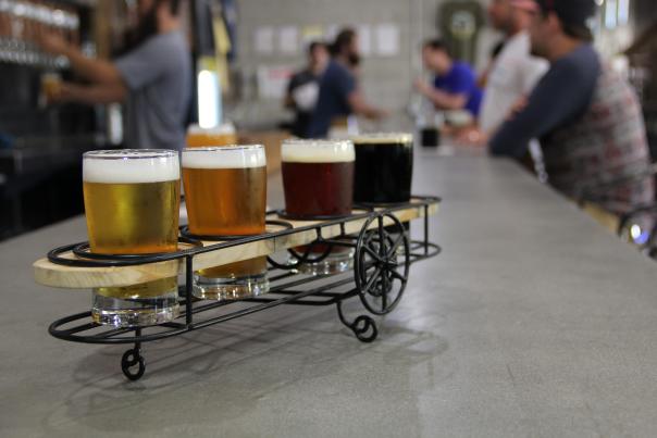 A flight of beer samples displayed apropros in wire-framed bi-plane-shaped holder at Crooked Can Brewing. Flight sits on bar with people in background.