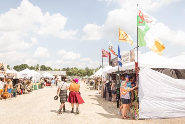 couple walking on dirt grounds, dressed in kilts between lined up vendors in white tents selling goods related to the Michigan Celtic Festival. Blue skies with white clouds, trees in distant background.