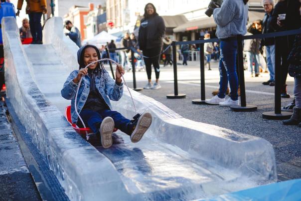Young girl on a sled sliding down an ice slide at an outdoor event.