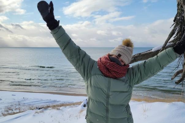 woman expresses joy while standing on snow covered hill overlooking lake michigan. she wears white snowpants, mint winter coat, burgundy scarf around her face and white knit hat.