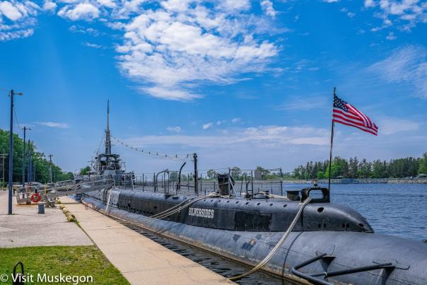 ww2 submarine docked in muskegon channel. a sidewalk runs along the channel, the sky is bright blue with some wispy white clouds. Green trees line the channel and an American flag flies on the stern of the ship.