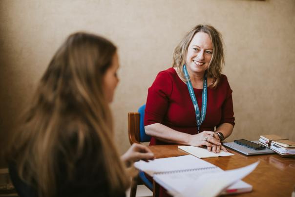 A woman sits at a desk and smiles at another woman out of focus