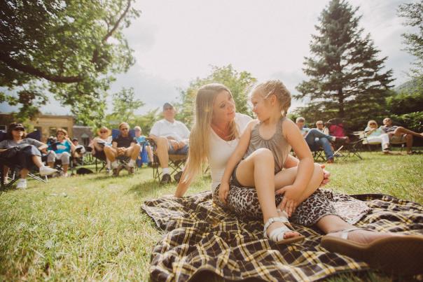 A girl and her mother sit on a blanket on the grass, smiling at each other with others sitting on lawn chairs.