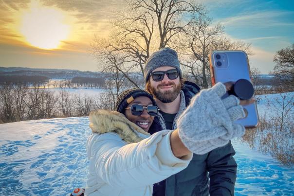A couple taking a selfie in the winter, surrounded by snow, with the Mississippi River and the sunset behind them.