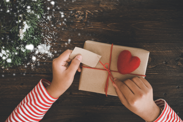 gift wrapped in brown paper tied up with red string and adorned with red felt heart sits on wooden table. someone is tying a the bow and adding tag. snow covered pine branches lay on table in upper left side of photo.