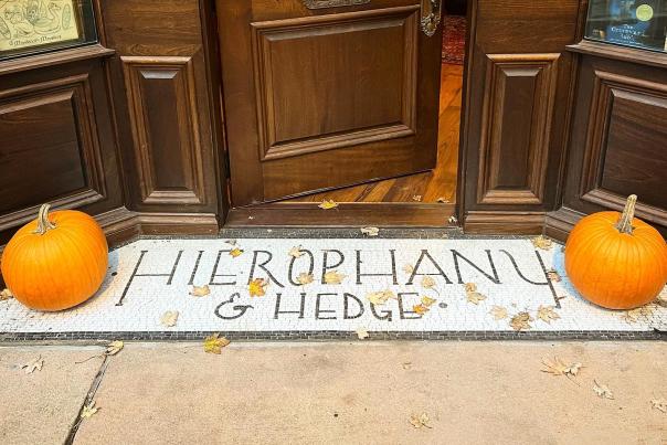 Image is of the bottom of the door and sidewalk up to the door that say's "Hierophany & Hedge" with a pumpkin on each side.