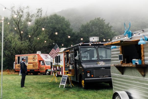 Four food trucks lined up in a grassy field, with a customer reading the menu for 'Betty's Biscuits.'