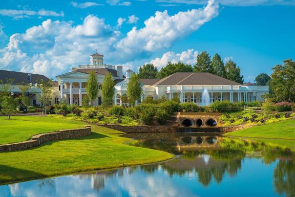 A scenic view of Huntsville Botanical Garden showcases the Eloise McDonald Propst Guest Center reflected in a tranquil pond. Lush landscaping, a small waterfall, manicured lawns, and vibrant greenery surround the elegant white building beneath a bright blue sky filled with puffy clouds.