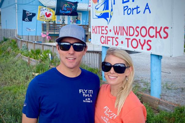 A couple in sunglasses pose together in front of a store called "Fly It! Port A"