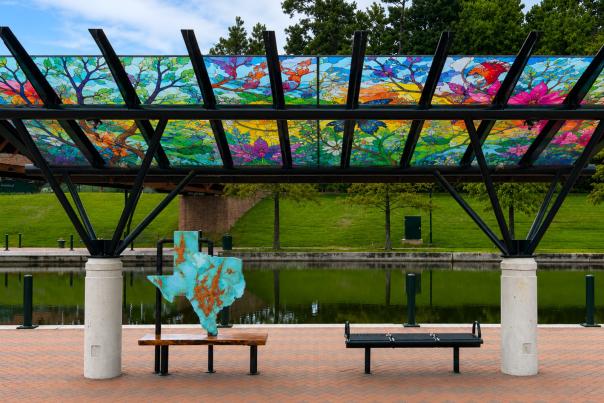 A rendering of the new trolley shelters with new digitally printed stained glass images of nature. This trolley stop resides beside The Woodlands Waterway, shielding The State of Texas art bench (a large, turquoise, Texas-shaped slap with a solid wooden bench).