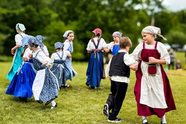 a group of children in period finnish american attire dance on a green lawn