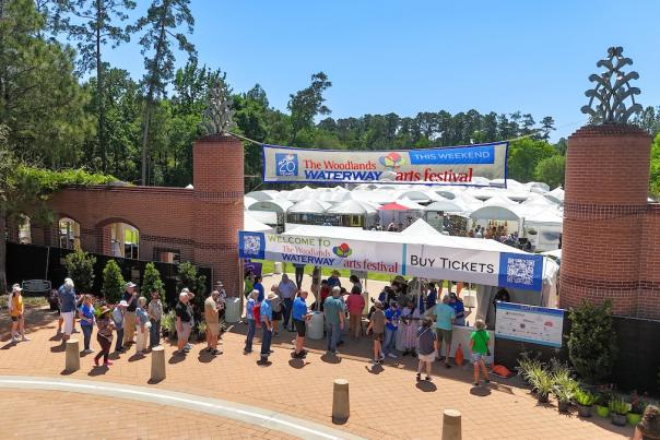 An elevated view of The Woodlands Waterway Arts Festival from the entrance to Town Green Park. People are lined up across the frame to buy tickets at the entrance to a park full of white tents.